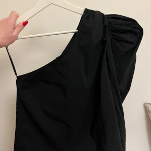 Svart oneshoulder blus - Snygg svart oneshoulder blus med en elegant och stilren design. Perfekt för festliga tillfällen. 