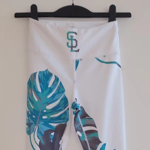 Vita leggings med bladmönster från Strong Lifestyle - Snygga vita leggings från Strong Lifestyle i storlek XS. De har ett coolt bladmönster i grönt och blått. Perfekta för träning eller en avslappnad dag. Materialet är 73% polyester och 27% spandex, vilket gör dem stretchiga och bekväma. Tvätta kallt och undvik torktumling för att bevara trycket.