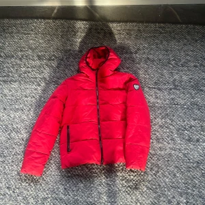 Röd pufferjacka från Emporio Armani - Säljer en snygg röd pufferjacka från Emporio Armani. Jackan har en dragkedja framtill och en huva för extra värme. Perfekt för kyliga dagar och har en stilren design med märkets logga på ärmen. Passar både till vardags och lite finare tillfällen.