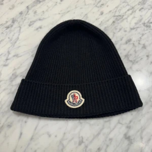 Moncler mössa - Säljer en snygg svart stickad mössa från Moncler. Den är i nytt skick och har en broderad logga framtill. Perfekt för vintern och håller dig varm med stil. Passar både till vardags och mer uppklädda tillfällen. Har kvitto 