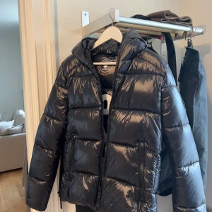 Svart pufferjacka från Save the Duck - Säljer en snygg svart pufferjacka från Save the Duck. Jackan är i mycket bra skick och perfekt för vintern. Den har en glansig yta och är utrustad med dragkedja och huva. Perfekt för att hålla dig varm och stilren under kalla dagar! 🖤