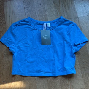 Blå croppad t-shirt från H&M - Säljer en snygg blå croppad t-shirt från H&M i storlek M. Den är gjord av 25% återvunnen bomull och har en enkel, stilren design. Perfekt för sommardagar eller som en basplagg i garderoben. Helt ny med tag kvar!