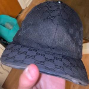 Svart Gucci Canvas Keps (Äkta) - Säljer en snygg svart keps från Gucci med deras ikoniska mönster (Canvas) . Kepsen har en klassisk design med en röd och grön randdetalj på sidan. Perfekt för att ge din outfit en lyxig touch. Passar både till vardags och fest! Priset kan diskuteras! MVH!