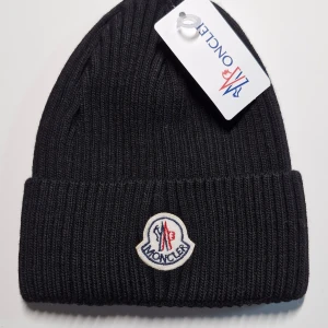 Svart ribbad mössa från Moncler - Säljer en snygg svart ribbad mössa från Moncler. Den är i nytt skick och har en broderad logga framtill. Perfekt för kalla vinterdagar och ger en stilren look. Passar de flesta tack vare one size-storleken.