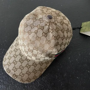 Beige keps från Gucci - Snygg beige keps från Gucci med deras ikoniska mönster i brunt. Kepsen har en justerbar rem baktill och en metallknapp på toppen. Perfekt för att ge din outfit en lyxig touch! Obs box och påse finns också. Byten går också 