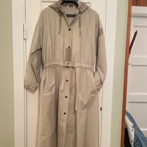 Beige sommarkappa från Luhta - Säljer en beige tunn men vindtät sommarkappa  från Luhta i mycket bra skick. Den har en huva och tryckknappar framtill samt ett knytband i midjan för en justerbar passform. 116 cm lång Perfekt för vår och höst! 🧥