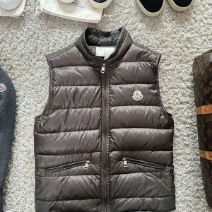 Brun dunväst från Moncler - Säljer en snygg brun dunväst från Moncler i bra skick. I storleken 2. Perfekt när du vill hålla dig varm men ändå se stilren ut. Det finns en liten fläck på framsidan, men den är knappt märkbar. Passar perfekt för både vardag och festliga tillfällen. Såklart är västen äkta och boxen samt påse kommer med, Skriv vid minsta lilla intresse