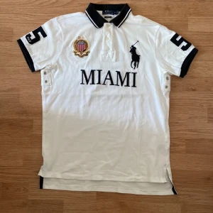 Vit pikétröja med Miami-tryck  - Säljer en snygg vit pikétröja från Ralph Lauren med svart Miami-tryck och nummer 5 på både ärmar och rygg. Tröjan har korta ärmar och en klassisk krage med knappar. Perfekt för en sportig och stilren look!