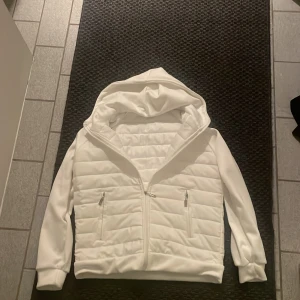 Vit cardigan  - Säljer en snygg vit cardigan jacka med huva. Den har dragkedja framtill och två fickor med dragkedjor. Perfekt för kyliga dagar och ger en stilren look. Jackan är långärmad och i mycket bra skick. Passar perfekt för höst och vår. Fick i julklapp föra året för tyvärr för liten för mig.