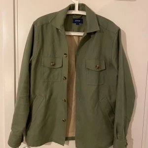 Grön jacka från Eton - Säljer en stilren grön jacka från Eton i storlek S. Jackan har långa ärmar och knappar framtill. Perfekt för vår och höst med sin klassiska design och praktiska fickor. Passar både till vardags och lite finare tillfällen. 🌿