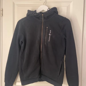 Svart huvtröja från Sail Racing - Säljer en svart huvtröja från Sail Racing i bra skick. Den har en dragkedja framtill och en broderad logga på bröstet och huvan. med långa ärmar och en skön passform.  Ordinarie pris runt 1000 kr