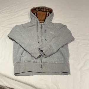Burrberry kofta - Säljer en snygg grå hoodie från Burberry med klassiskt rutigt mönster i luvan. Den har dragkedja framtill och är i mycket bra skick. Perfekt för höst och vår när du vill vara både bekväm och stilren. Passar till alla tillfällen! 💫 storleken är S men passar defentivt Xs dragkedjan är trög!!!