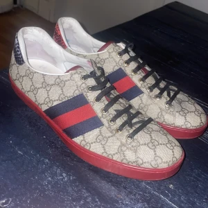 Gucci sneakers med röd och blå rand - Säljer ett par snygga Gucci sneakers i beige med klassiskt mönster och röd-blå rand. Skorna har en röd sula och detaljer i rött och blått ormskinnsmönster på hälen. Perfekta för att ge din outfit en lyxig touch! Använda men i bra skick.