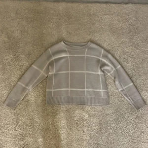 Grå rutig stickad tröja - Säljer en grå  beige stickad tröja med rutigt mönster. Tröjan har långa ärmar och ribbade kanter vid ärmslut och nederkant. Perfekt för höst och vinter! Har också 2 hål men syns knappast på. Står inte vart den är köpt men själv köpte jag på second hand!