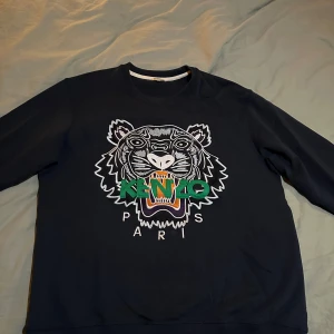 Mörkblå sweatshirt från Kenzo - Säljer en svart sweatshirt från Kenzo med ett coolt tigerbroderi i vitt, grönt och orange på framsidan. Tröjan har långa ärmar och en rund halsringning. Perfekt för en avslappnad stil eller att sticka ut lite extra. Passar både till vardags och fest!