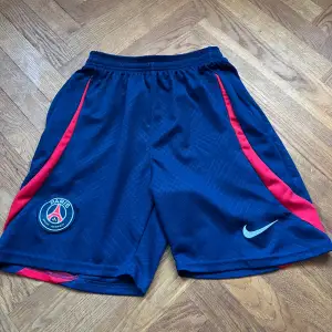 Säljer ett par snygga PSG shorts från Nike i blått med röda detaljer. Perfekta för fotbollsträningen eller bara för att chilla. De har en elastisk midja för bästa passform och är gjorda i ett lätt och bekvämt material. Fickor med dragkedja, ibland hoppar dragkedjan ut men går att sätt tillbaka snabbt. Hör av dig vid frågor👋