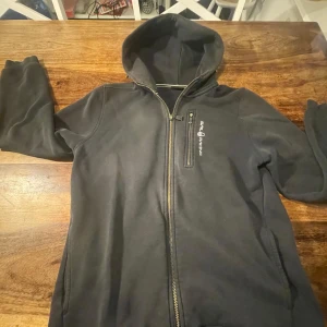 Svart hoodie från Sail Racing - Säljer en svart hoodie från Sail Racing i bra skick. Den har en dragkedja framtill och en praktisk huva. Perfekt för höst och vår, med en stilren design och bekvämt material. Passar både till vardags och lite sportigare aktiviteter.