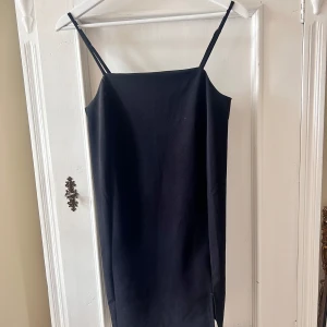 Svart klänning med smala axelband - Säljer en stilren svart klänning med smala axelband. Den har en enkel och elegant design med en liten slits på sidan. Perfekt för både vardag och festliga tillfällen. Klänningen är i ett mjukt och bekvämt material.