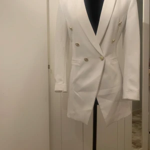 Vit kavaj med guldknappar - Säljer en elegant vit kavaj med dubbelknäppning och stiliga guldknappar med medföljande byxor. Kavajen har en klassisk passform och är perfekt för både formella och festliga tillfällen. 