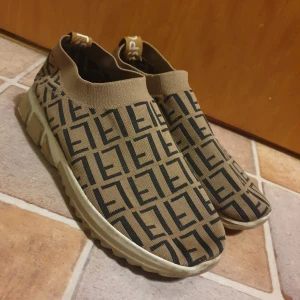 Beige och svarta sneakers med mönster - Säljer ett par snygga sneakers i beige och svart. De är i nyskick och har en bekväm passform. Perfekta för både vardag och fest! Skorna har en elastisk öppning för enkel på- och avtagning. Obs: flärpen har börjat lossna på ena skon, se sista bilden. 