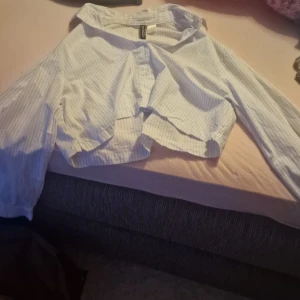 Vit och blå randig croppad skjorta från H&M Divided - Säljer en superfin vit och blå randig croppad skjorta från H&M Divided. Den har långa ärmar och knappar framtill. Perfekt för en avslappnad stil eller att klä upp med accessoarer. Passar perfekt till vår och sommar!
