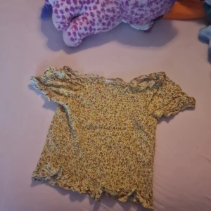 Blommig topp från Lindex - Säljer en söt blommig topp från Lindex i storlek 152. Den har korta ärmar och ett gulligt volangmönster längs kanterna. Perfekt för sommarens varma dagar!