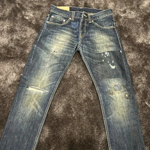 Blå jeans med slitningar - Säljer ett par snygga blå jeans från Dondup. De har en cool, sliten look med flera distinkta slitningar och en vintage känsla. Jeansen har en klassisk femficksdesign och är perfekta för en avslappnad stil. Passar bra till både vardag och fest!