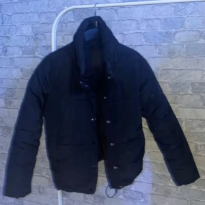 Svart dunjacka - Säljer en svart dunjacka som är perfekt för kalla dagar. Jackan har en klassisk design med långa ärmar och knappar framtill. Den är varm och bekväm, idealisk för både höst och vinter.