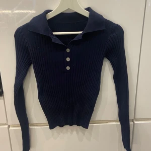 Mörkblå ribbad tröja med krage - Säljer en snygg mörkblå ribbad tröja med krage och knappar framtill. Tröjan har långa ärmar och en tight passform, perfekt för höst och vinter. Den är både stilren och bekväm, idealisk för både vardag och fest! 