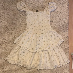 Blommig sommarklänning med volanger - Säljer en superfin vit klänning med små gula blommor. Den har en smockad överdel och söta volanger på kjolen. Klänningen är ärmlös med breda axelband och perfekt för sommardagar. Materialet känns lätt och luftigt, perfekt för varma dagar!