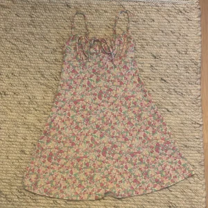 Blommig sommarklänning med knytband - Säljer en superfin blommig sommarklänning med tunna axelband och knytband framtill. Klänningen har ett härligt mönster i rosa, grönt och gult som verkligen ger sommarkänsla. Perfekt för varma dagar eller en mysig picknick i parken!