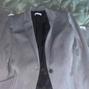 Grå kavaj från Mango - Säljer en stilren grå kavaj från Mango i storlek S. Kavajen har en klassisk design med knappar framtill och ett snyggt knytband i ryggen för en justerbar passform. Perfekt för både kontoret och festligare tillfällen!
