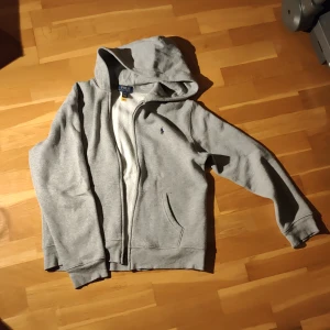 Grå hoodie från Ralph Lauren - Säljer en stilren grå hoodie från Ralph Lauren med dragkedja och broderad logga på bröstet. Perfekt för en avslappnad look. Den har en mjuk insida och praktiska fickor framtill. Str: 160 (14/15år)