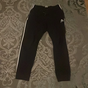 Svarta mjukisbyxor från Adidas - Säljer ett par svarta mjukisbyxor från Adidas med vita ränder längs sidorna och logga på benet. Byxorna har en elastisk midja och är perfekta för en avslappnad stil.
