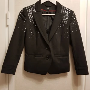 Mörkgrå tre kvarts arm, kavaj med glittret på axlarna strl 38 från H&M - Snygg mörkgrå kavaj från H&M med glitter på axlarna. Kavajen har en figursyd passform med två knappar framtill och två fikor. Perfekt för att ge din outfit en edgy touch. Någon av glutter sakerna kan ha trillar av men dom sitter bra skivk är mycket bra