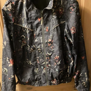 Svart mönstrad jacka från Odd Molly - Säljer en svart jacka från Odd Molly med blommigt mönster och dragkedja framtill. Jackan har långa ärmar och en cool design med små detaljer i olika färger. Perfekt för att ge din outfit en unik touch!