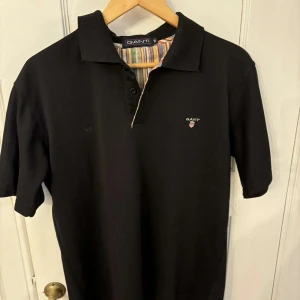 Svart pikétröja från GANT - Snygg svart pikétröja från GANT med klassisk krage och korta ärmar. Tröjan har en diskret logga på bröstet och en färgglad randig insida av kragen. Perfekt för en stilren look!