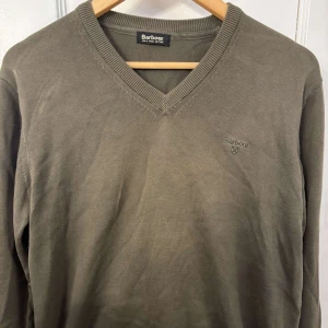 Olivgrön tröja från Barbour - Snygg olivgrön tröja från Barbour med v-ringning. Perfekt för en stilren och avslappnad look. Tröjan har ribbade kanter och en diskret broderad logga på bröstet.