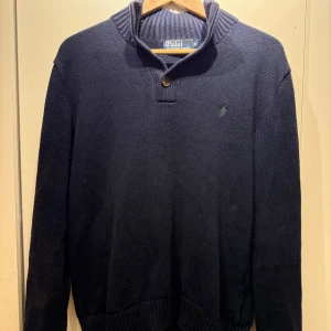 Mörkblå stickad tröja från Polo Ralph Lauren - Säljer en snygg mörkblå stickad tröja från Polo Ralph Lauren med en grön broderad logga på bröstet. Tröjan har en ribbad krage och knappdetalj vid halsen.
