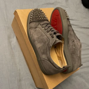 Christian Louboutin skor - Snygga grå sneakers i mocka från Christian Louboutin med ikoniska röda sulor. Skorna har nitar på tån och snörning framtill. Perfekta för att ge en edgy touch till din stil. Allt medkommer box , två dustbags, extra snören och kvitto. Skorna är köpta i Hollywood och cw:n finns inte längre.Skorna missar en spik men finns att köpa.