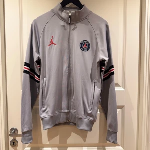 Grå och svart träningsjacka och byxor från Jordan - Set: Snygg grå träningsjacka med dragkedja och PSG-logga, samt matchande svarta byxor med röd och vit randdetalj. Perfekt för sportiga tillfällen med stil. Jackan har långa ärmar och byxorna har elastisk midja för bekväm passform.