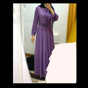 Lila festklänning  - Elegant lila festklänning med plisserade långärmar och knytband i halsen. Klänningen har bälte och osynlig dragkedja i midjan för en smickrande passform. Endast använd en gång. 