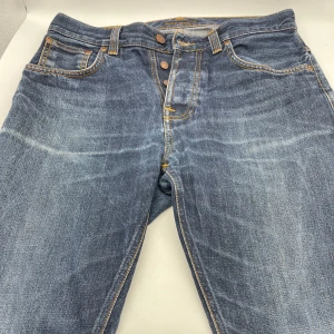 Blå jeans från Nudie Jeans (sleepy sixten) - Säljer ett par snygga blå jeans från Nudie Jeans. De har en klassisk femficksdesign med orange sömmar och knappar i gylfen. Mycket trendiga och tidslösa byxor som tyvärr är för små för mig