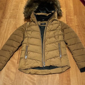 Brun jacka Everest - Snygg brun dunjacka från Everest med pälskantad huva och dragkedjor. Perfekt för kyliga dagar med sin vadderade design och långa ärmar. Jackan har praktiska fickor med dragkedja och en stilren look.