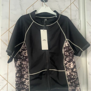 Svart kortärmad surftröja - Oanvänd svart kortärmad tröja med blommönster i vitt på sidorna. Tröjan har en dragkedja framtill. Bra utifall att man ska surfa och inte vill bränna axlarna. Baby tee style. Badkläders material.