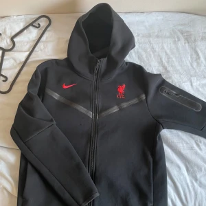 Svart hoodie med Liverpool FC-logga från Nike - Säljer en svart hoodie från Nike med Liverpool FC-logga i rött på bröstet. Hoodien har dragkedja och en praktisk ficka på ärmen. Skicket är 8/10 förutom en liten lagning som syns på bild fyra. Detta syns knappt när man har den på sig. Storlek L