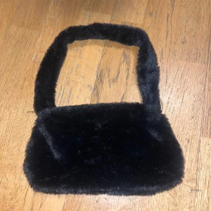 Svart pälsväska - Snygg och trendig svart axelväska i pälsmaterial. Perfekt för att ge din outfit en lyxig touch. Väskan har en mjuk och fluffig yta med ett bekvämt handtag.