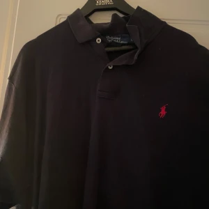 Svart pikétröja från Ralph Lauren - Klassisk svart pikétröja från Ralph Lauren med den ikoniska röda logotypen på bröstet. Tröjan har en krage med två knappar och korta ärmar. Perfekt för en stilren och avslappnad look.