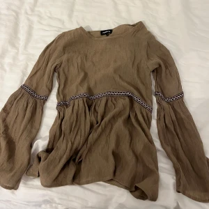 Beige brub långärmad topp  - Säljer en beige långärmad topp köpt i Spanien med dekorativa band i midjan och på ärmarna. Toppen är tillverkad i ett lätt material, perfekt för vår och sommar. Lite bohemisk vibe. 