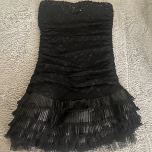 Svart axelbandslös klänning med spets och paljetter - Snygg svart axelbandslös klänning med spetsdetaljer och paljetter vid bysten. Klänningen har en volangkant i flera lager. Perfekt för nyår eller annan fest, tex maskerad. OBS KAN MÖTAS 31 DECEMBER om någon behöver akut nyårs klänning 